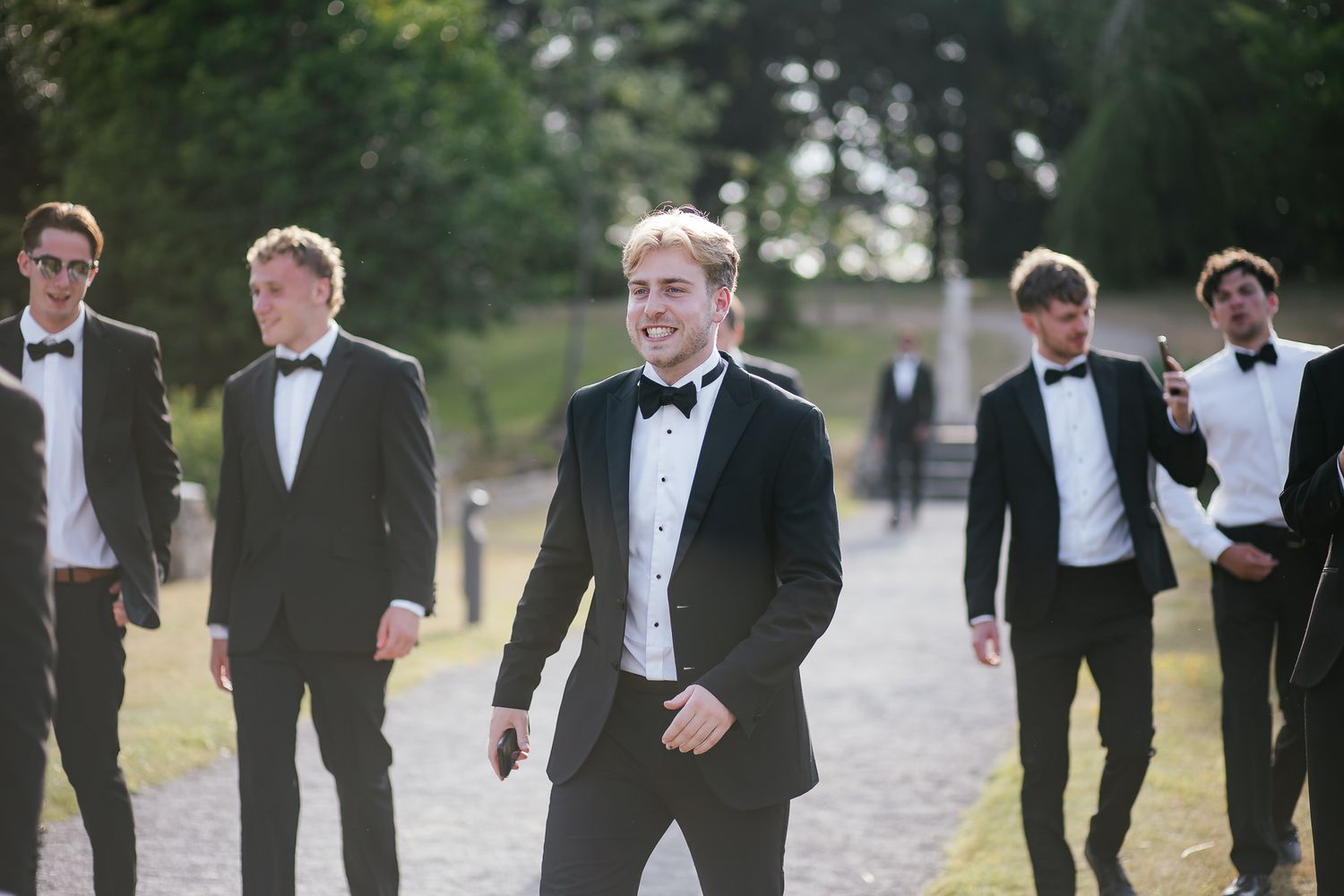 A group of groomsmen in black tuxedos walk along a path outdoors.