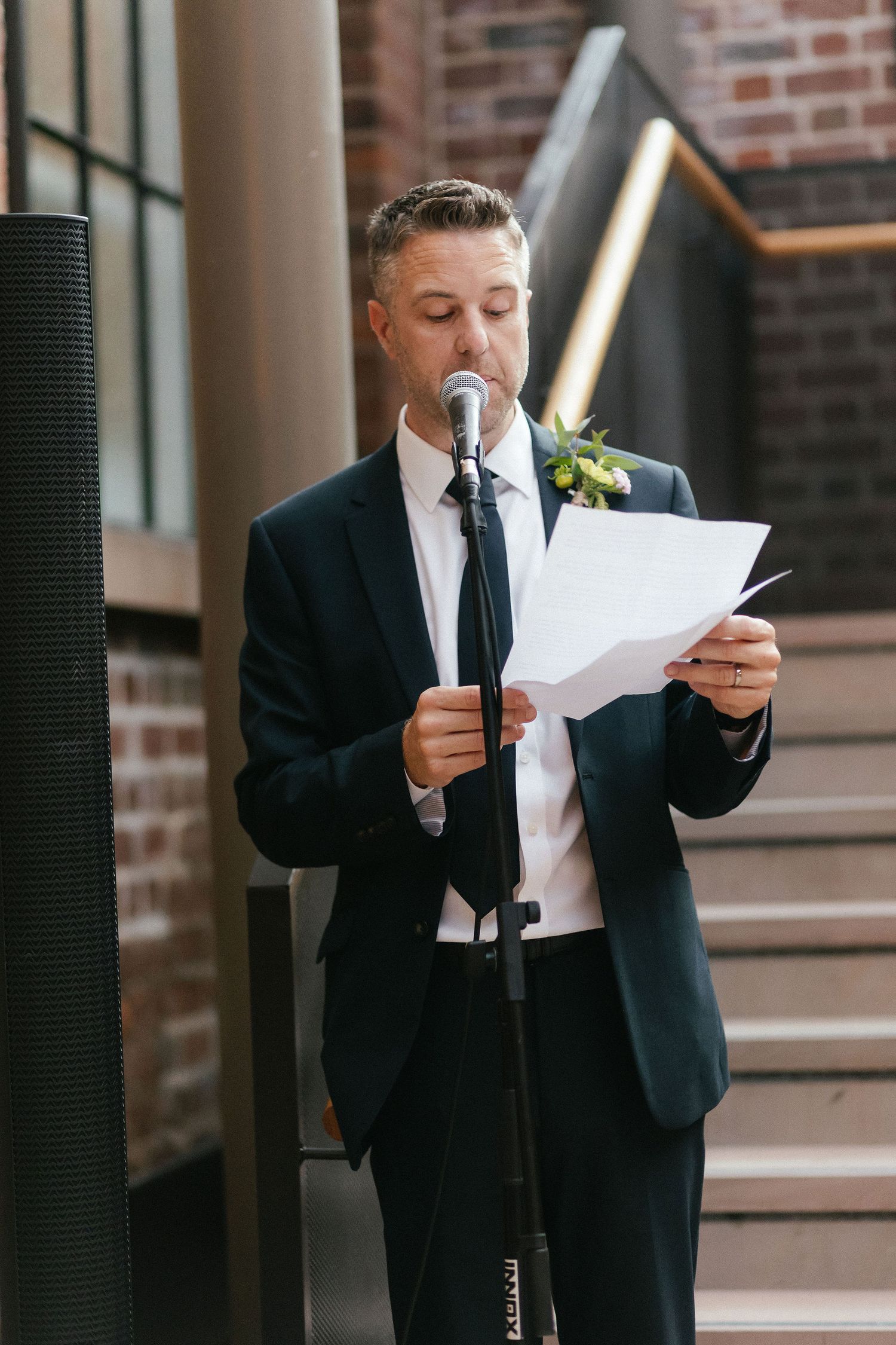 Man in dark suit with boutonniere reads from paper at microphone stand during wedding ceremony speech.