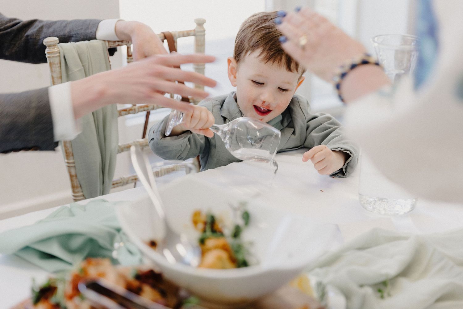 Adorable toddler in grey suit reaching for water bottle at wedding reception dinner table.