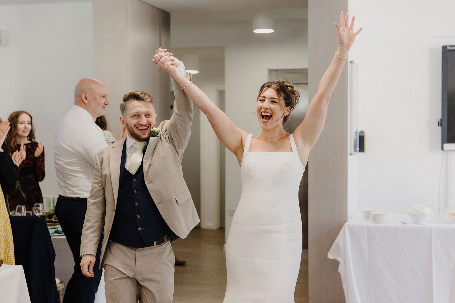 Joyful newlyweds raise their hands in celebration as they enter their wedding reception, bride in white gown and groom in tan suit.
