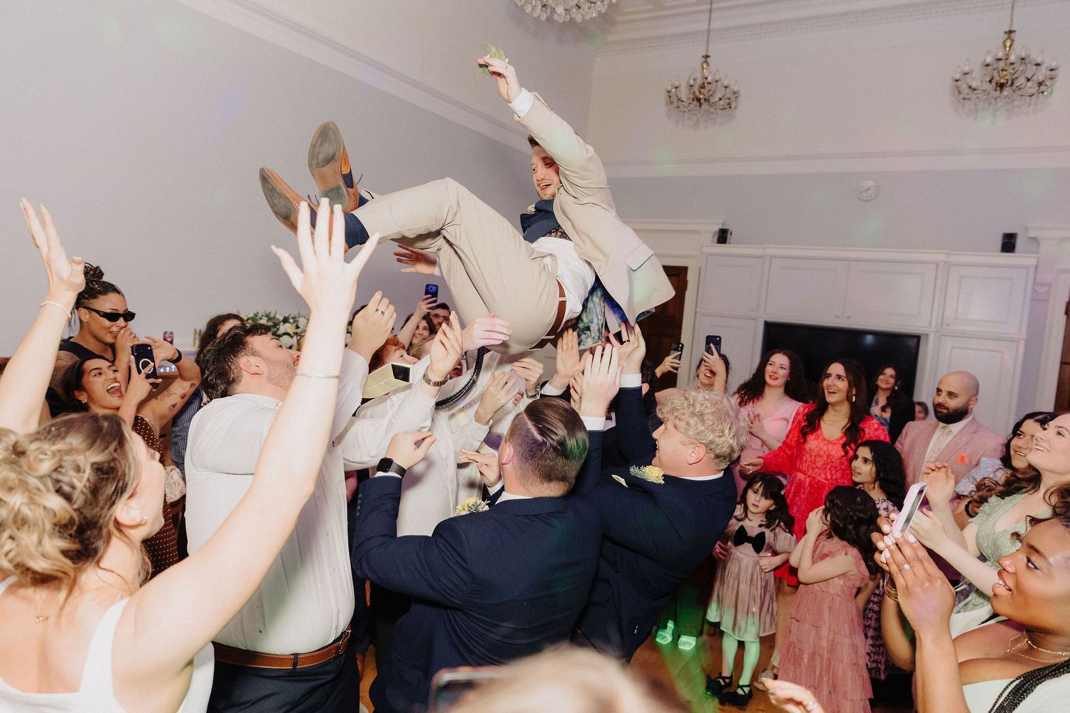 Guests crowd-surfing a man in a suit at a lively wedding reception dance floor celebration.
