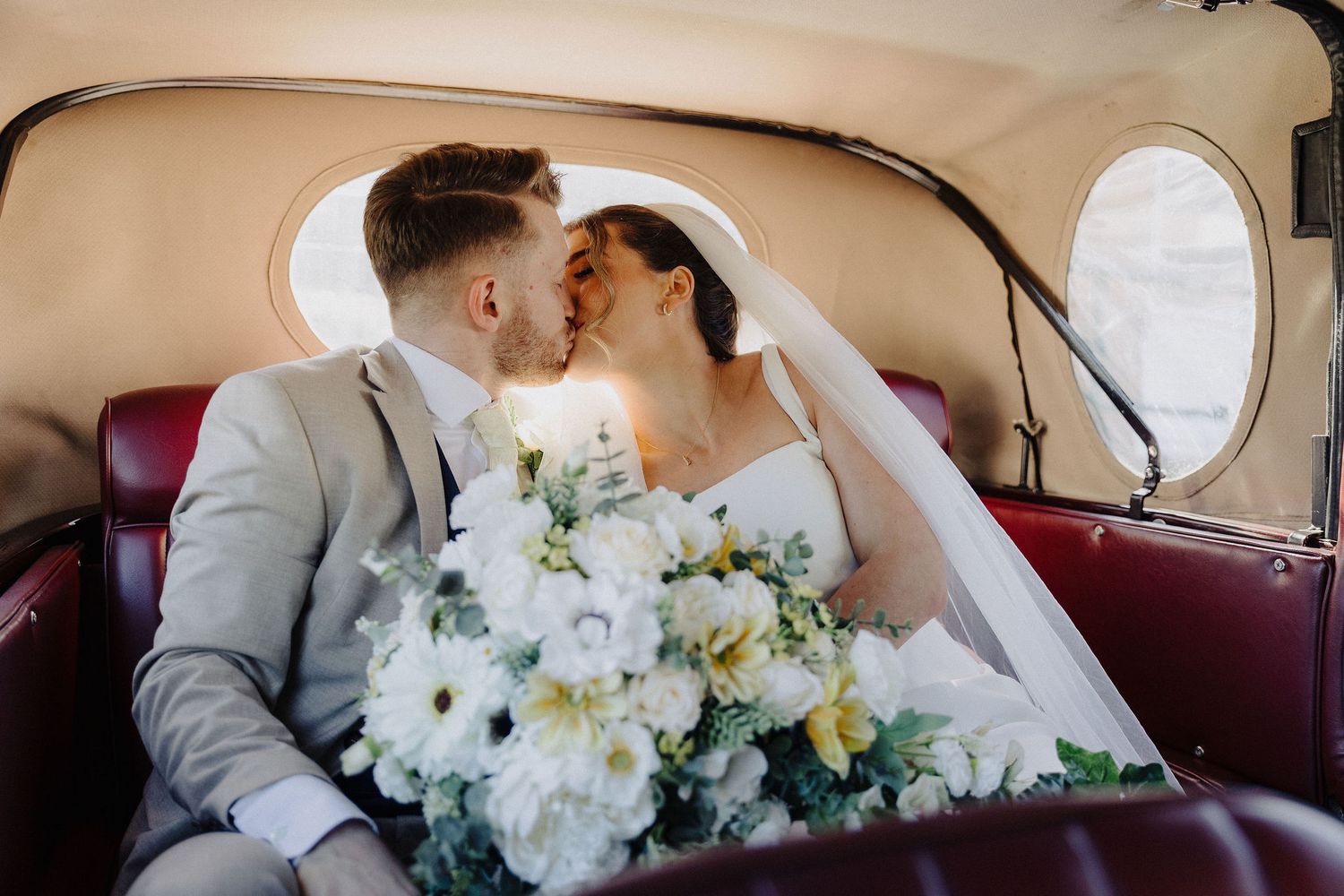 Bride and groom share a romantic kiss in a vintage car, bride holding a white floral bouquet.