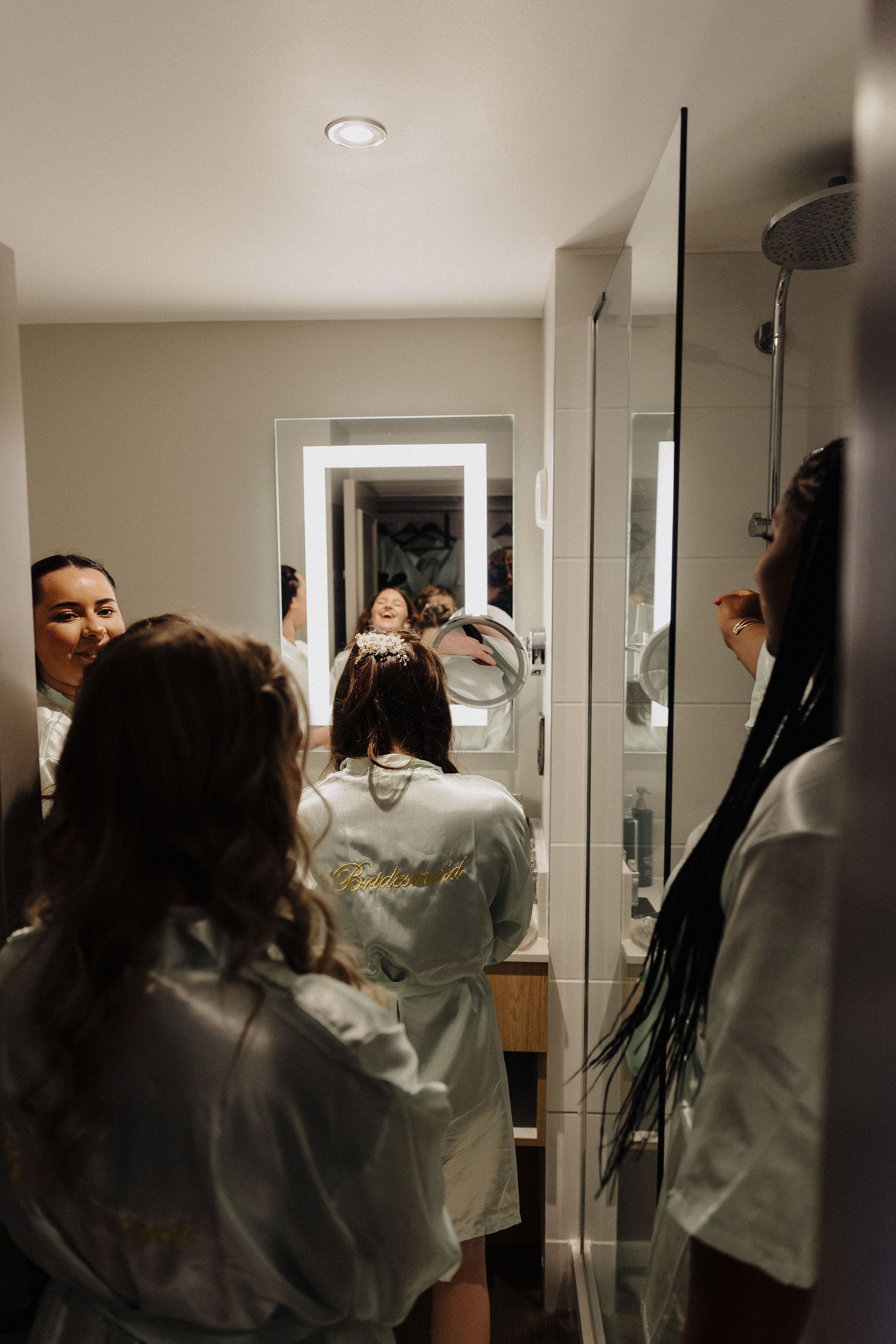Bridesmaids in white robes getting ready in a modern bathroom with LED mirror during bridal preparations.