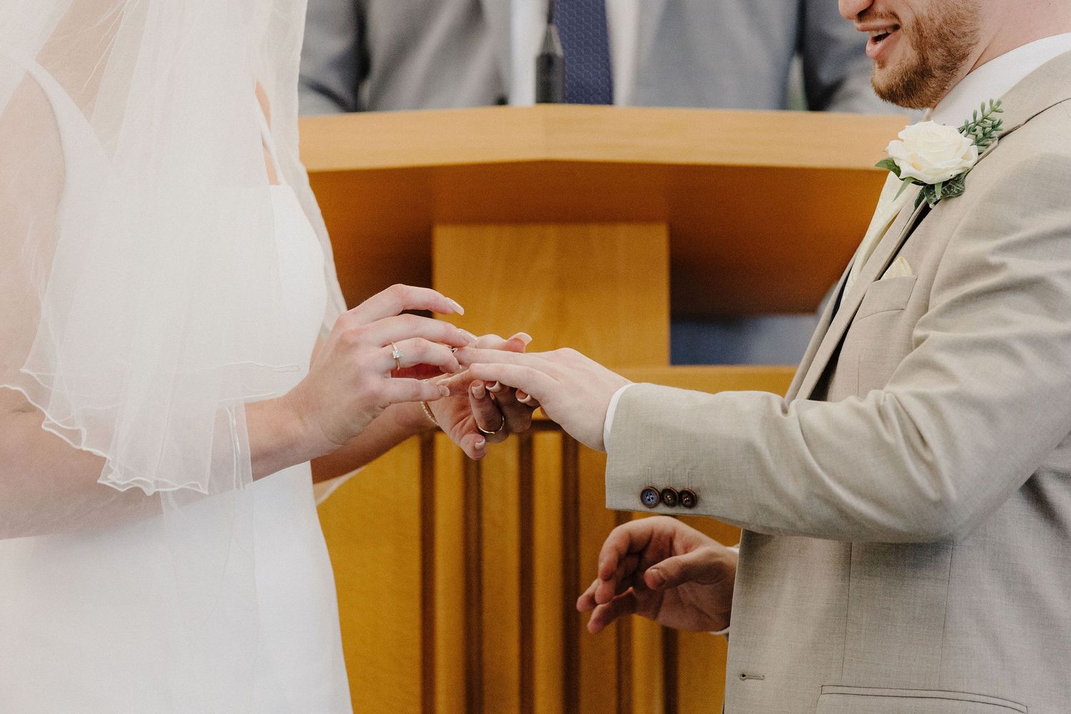Bride placing wedding ring on groom's finger during ceremony, groom wearing beige suit with white boutonniere.