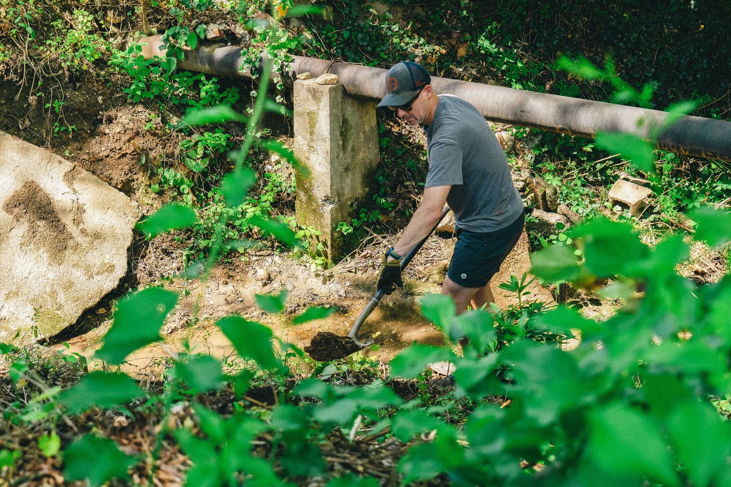 Man digging with a shovel in a lush green wooded area near a concrete post and large rock.