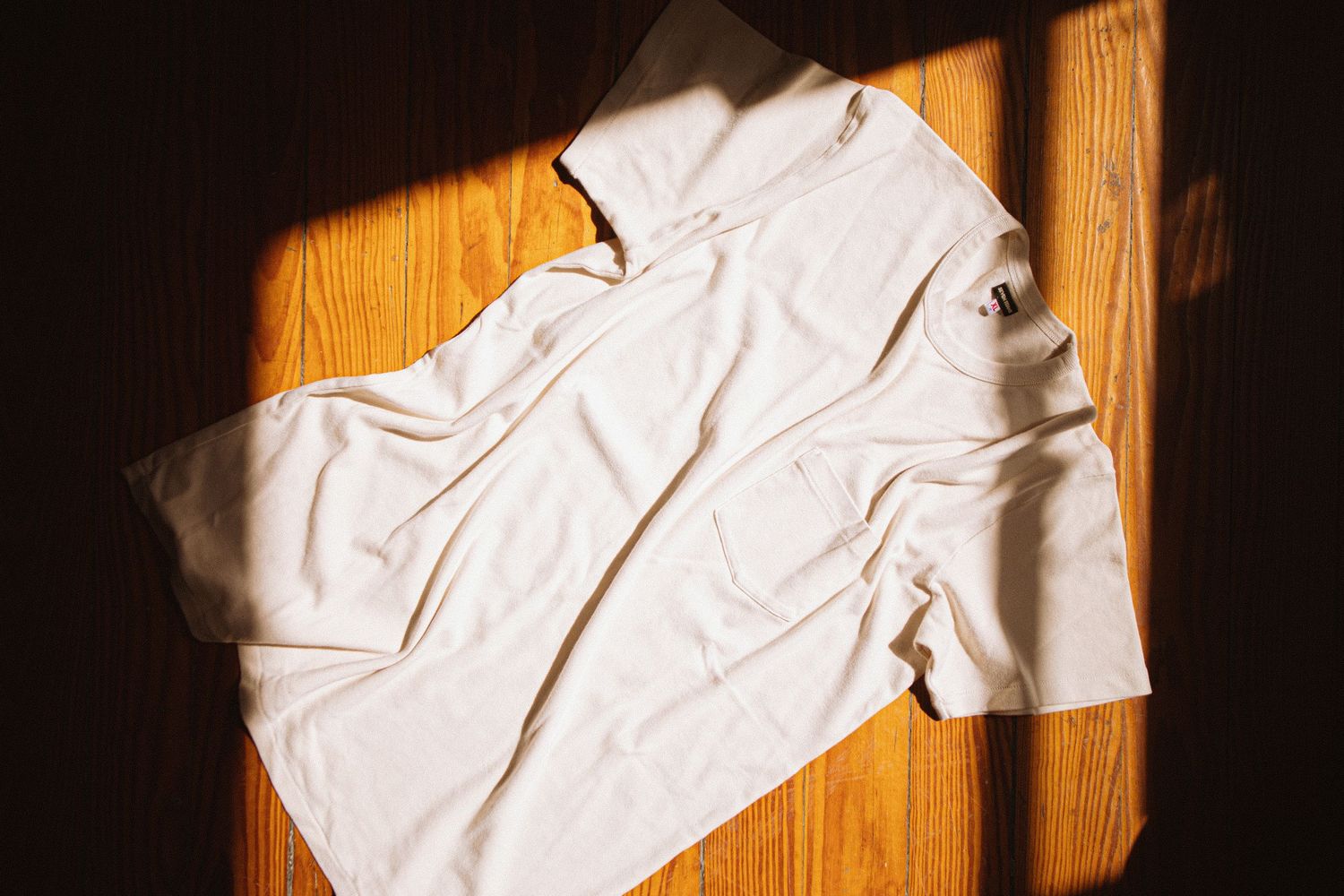A wrinkled white t-shirt laid flat on a wooden surface with dramatic sunlight casting shadows.