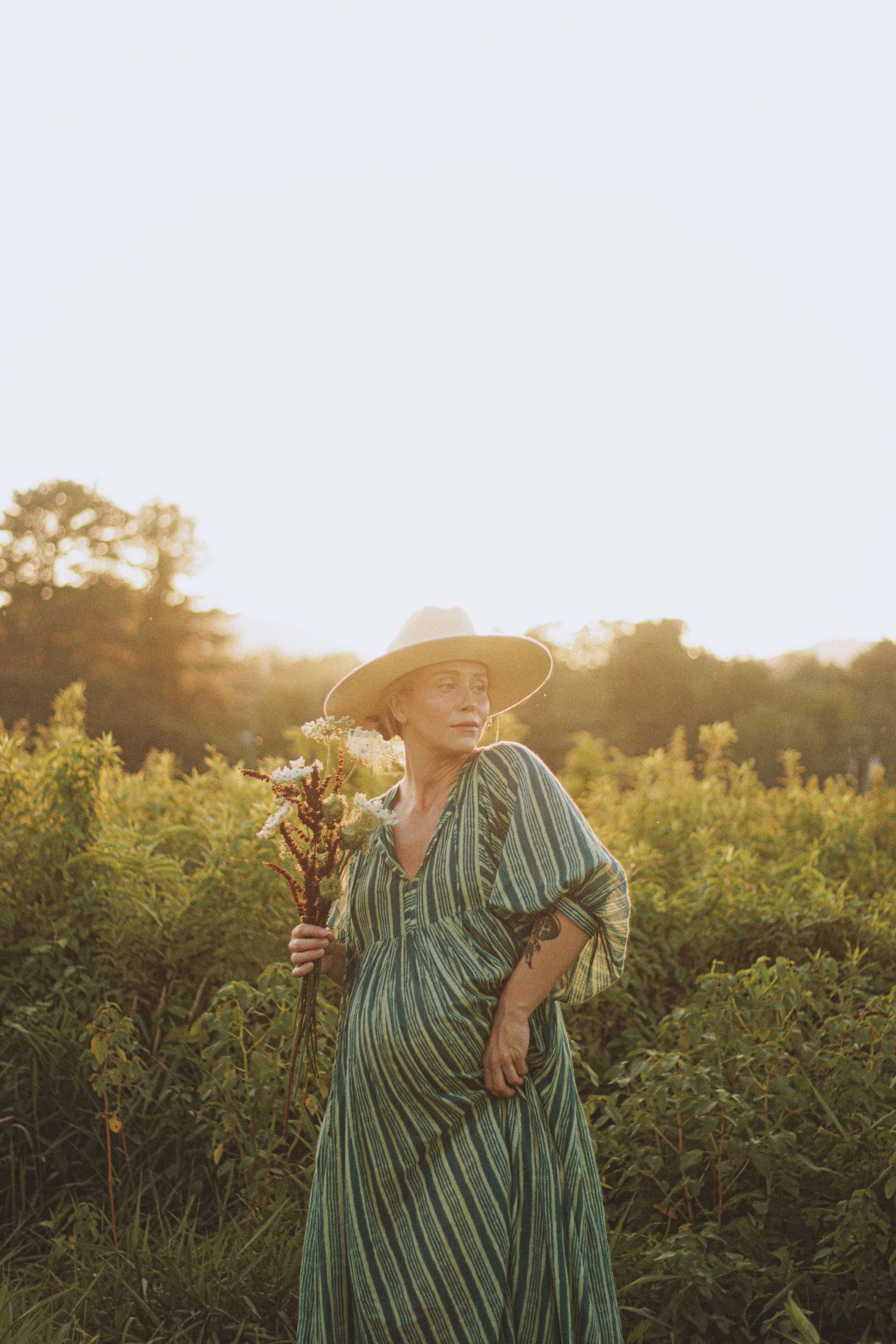 Woman in striped green dress and straw hat holding wildflowers in a sunlit field at golden hour.