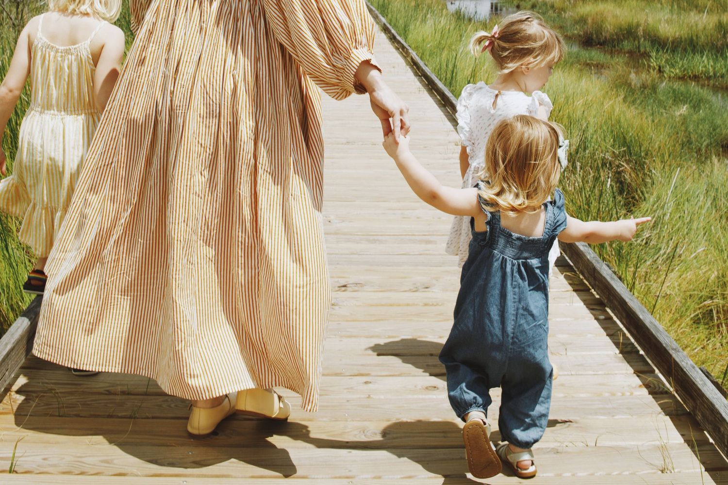 Mother walking on wooden boardwalk holding hands with two young children in summer outfits near tall grass.