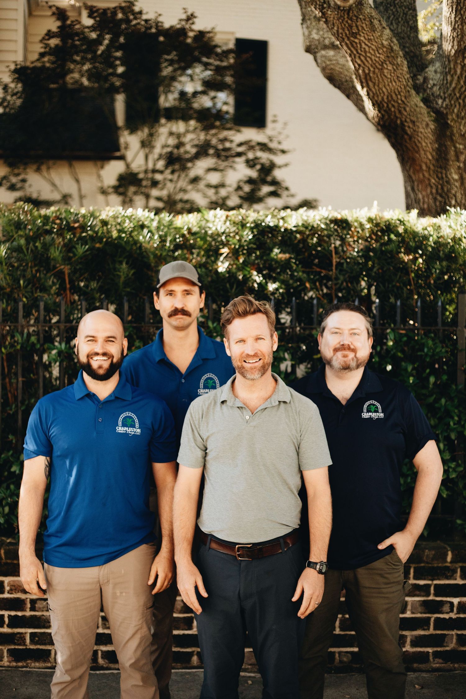 Four smiling men in blue polo shirts pose together outdoors near a brick wall and green hedges.