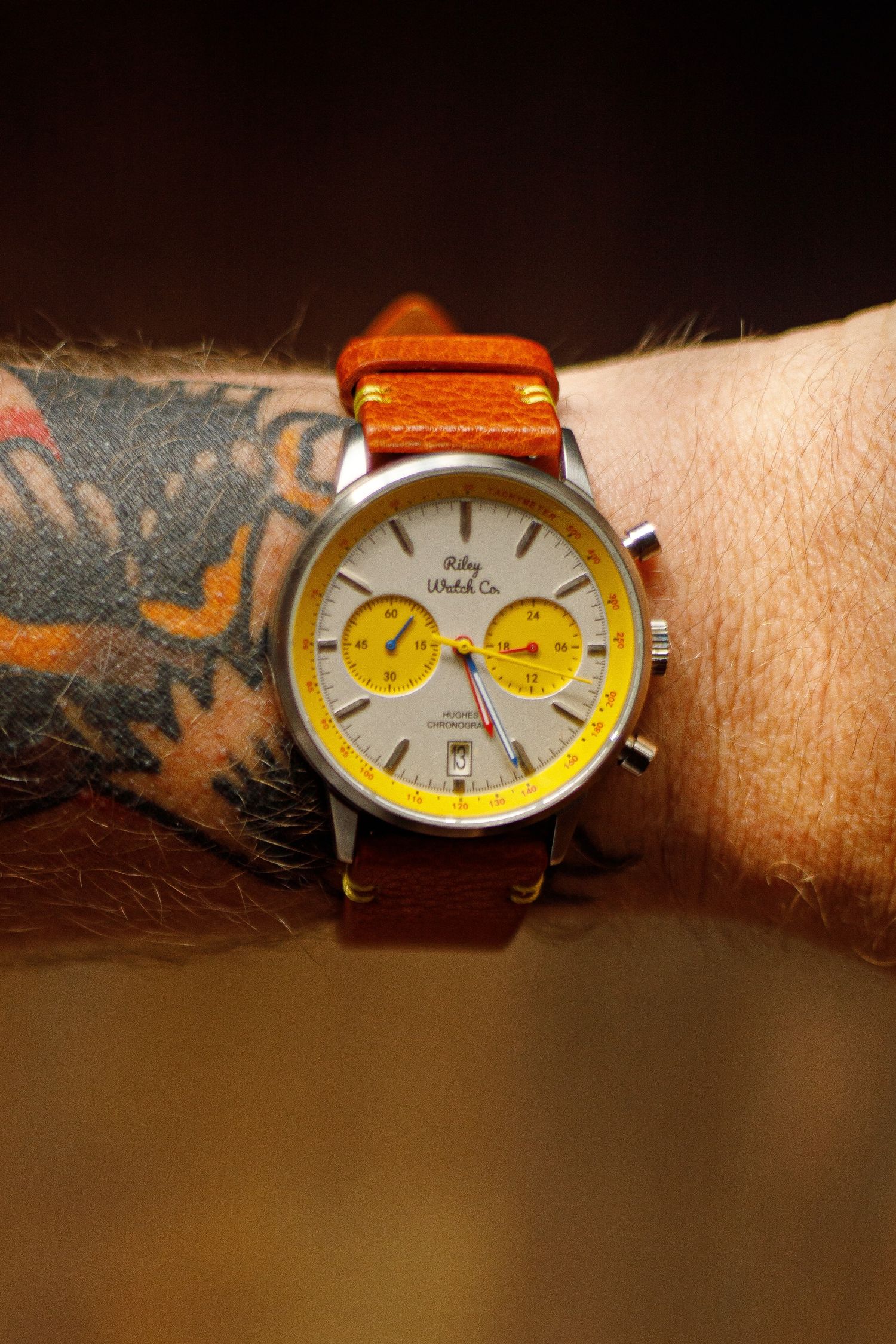 Riley Watch Co. chronograph with yellow subdials and a cream dial on a tan leather strap, worn on a tattooed wrist.