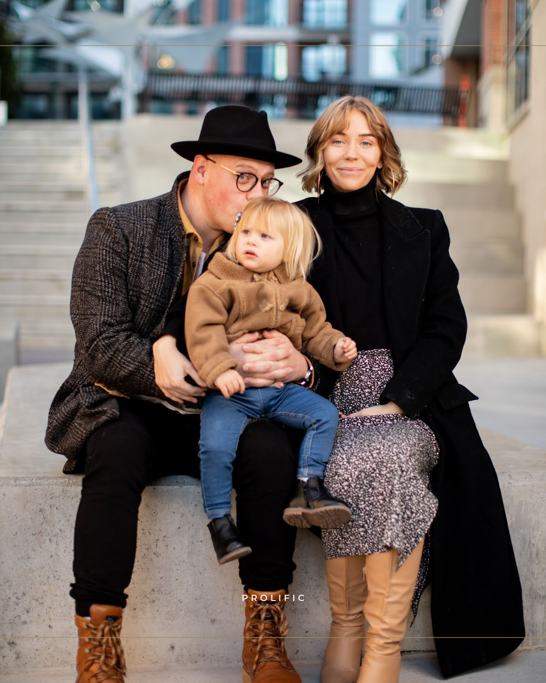 Family of three sitting on steps outdoors, parents in black coats with toddler in brown jacket.