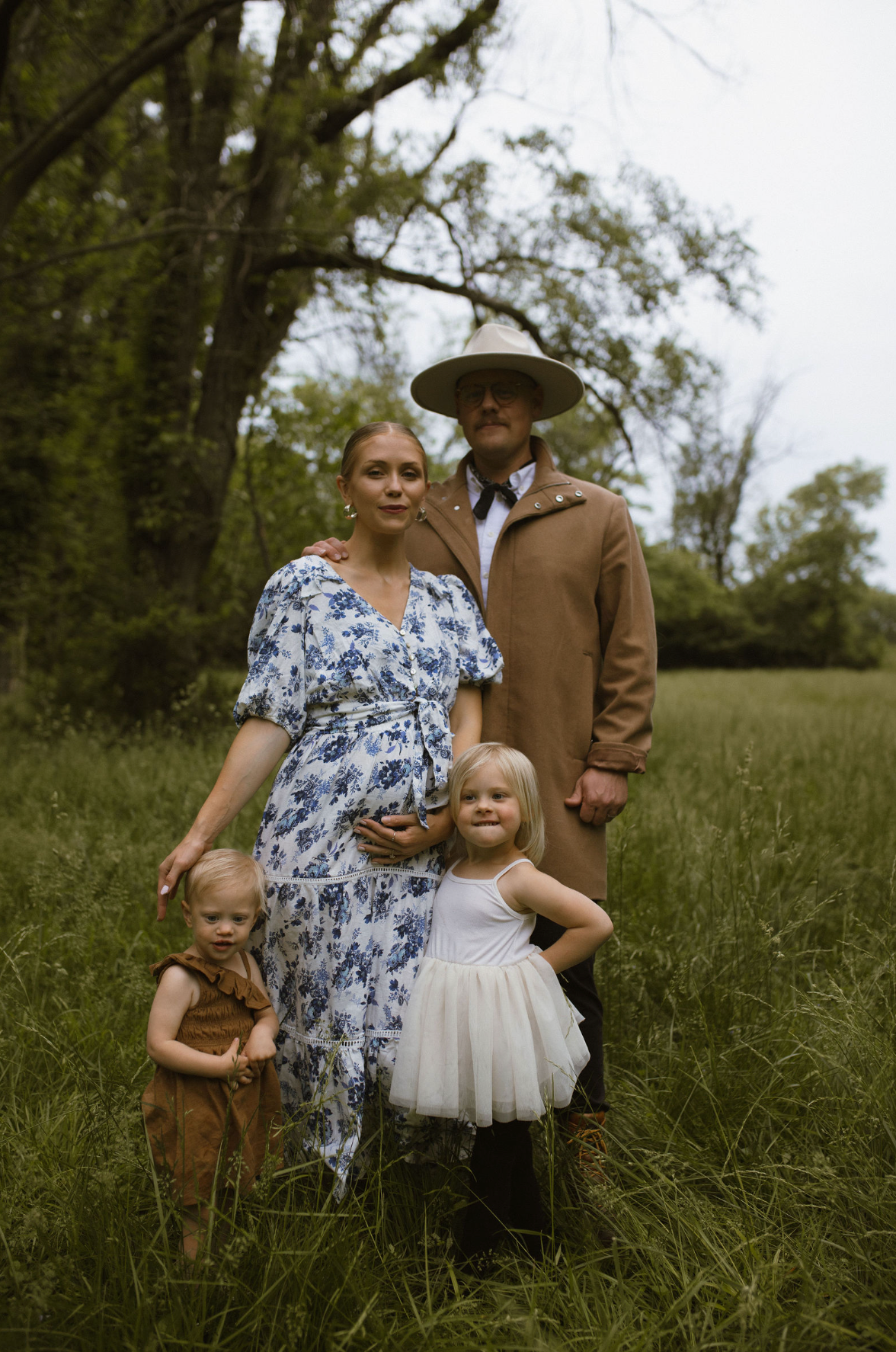 Family of four poses in a lush green field, mother in floral dress, father in tan coat and hat, two young children.