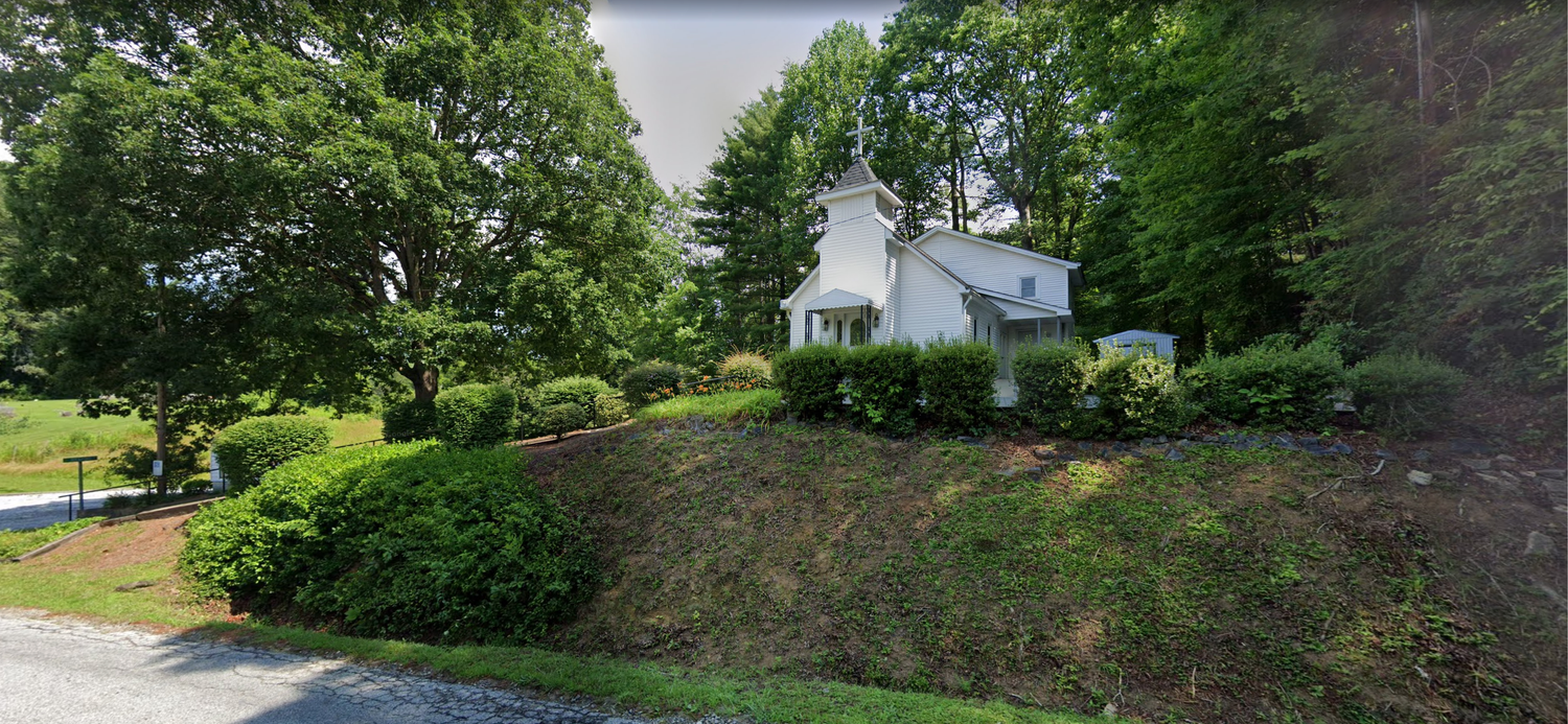 Small white church nestled among lush green trees on a hillside in a peaceful rural setting.
