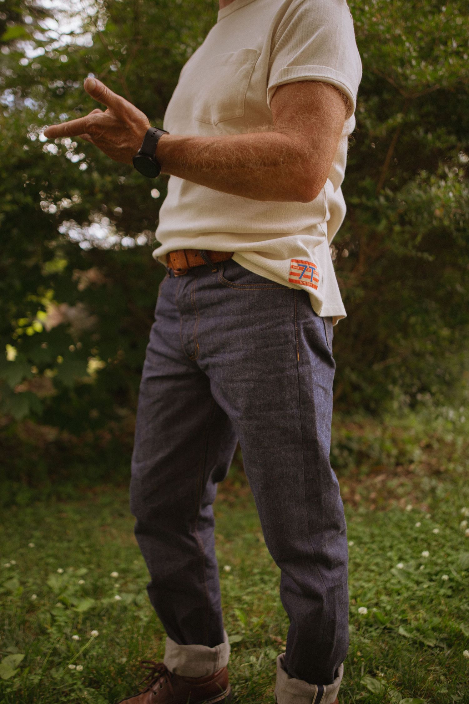 Man wearing cream t-shirt, dark selvedge denim jeans with cuffed hem, brown leather belt, and a watch outdoors.