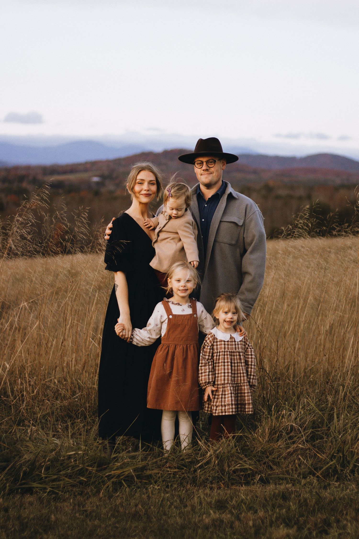 Family of five poses together in golden field with mountain range visible in background.