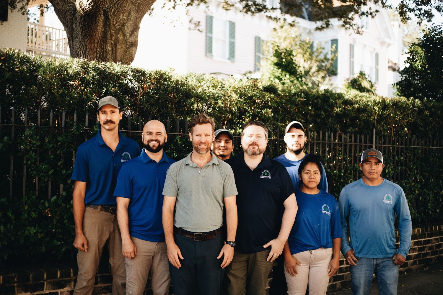 A group of seven team members in blue uniforms posing outdoors in front of a white building and green trees.