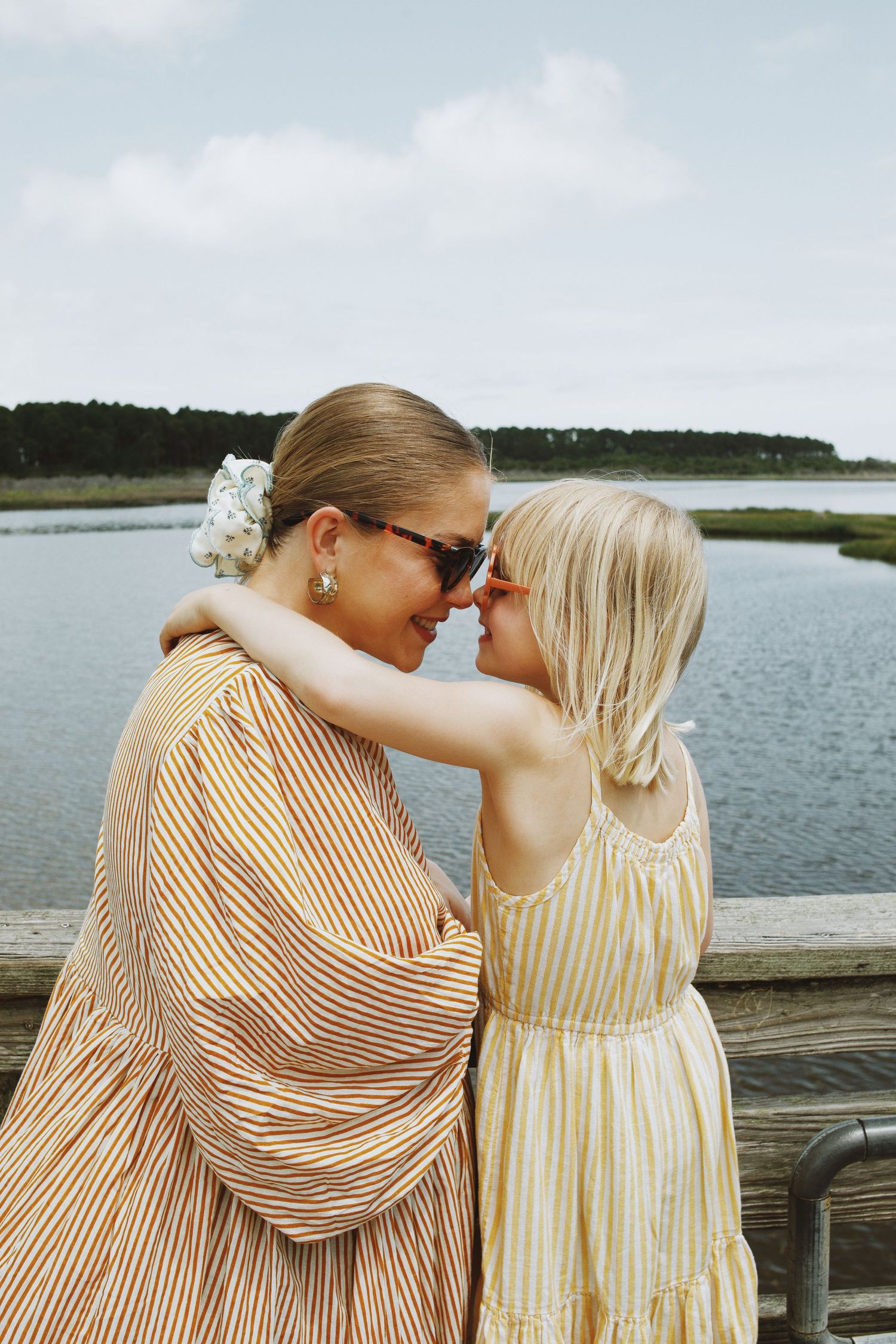Mother and daughter in matching striped dresses touch foreheads on a waterfront dock, sharing a tender moment.