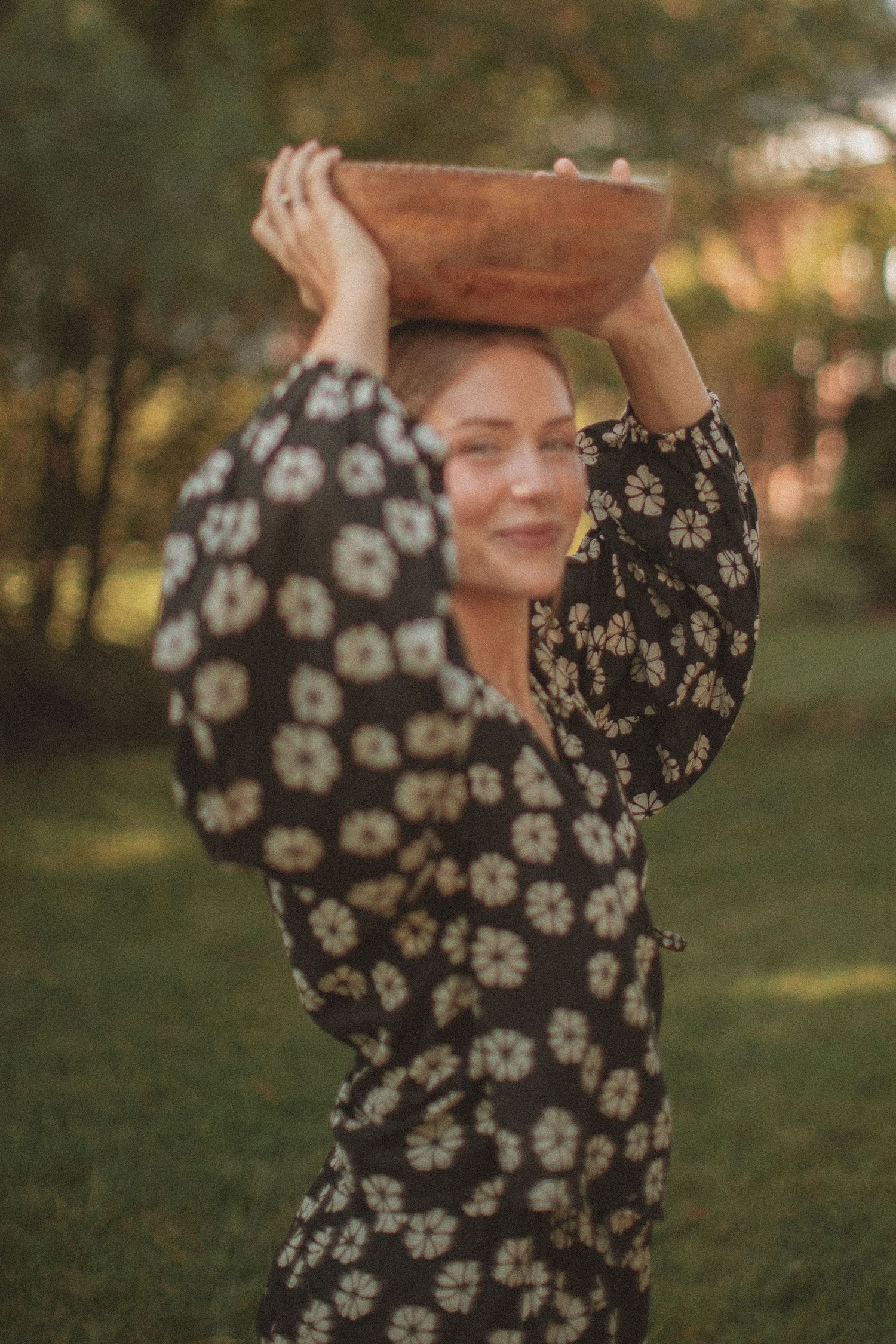 Woman in floral dress balancing a wooden bowl on her head outdoors in a garden setting.