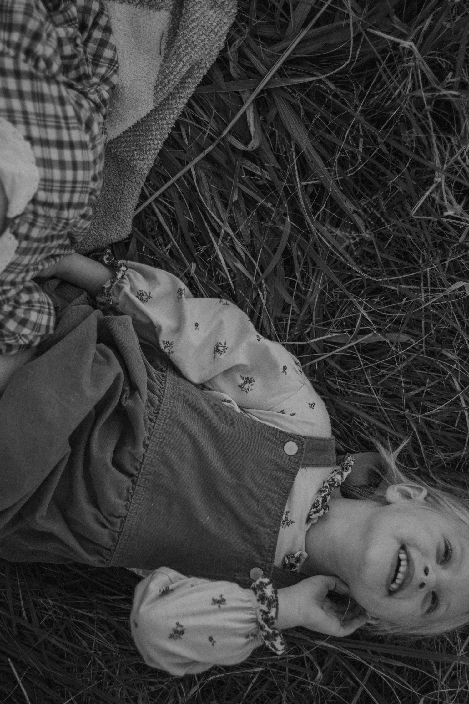 Black and white photo of a laughing child lying on grass, wearing overalls and a floral blouse.