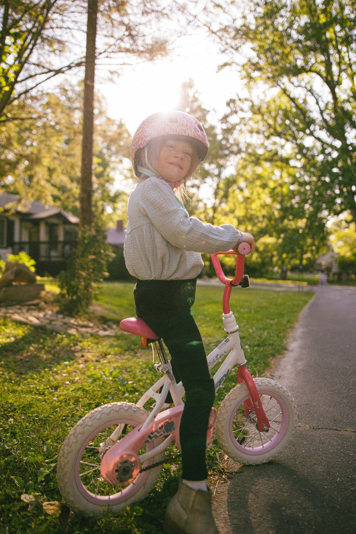 Young girl in pink helmet rides a small pink and white bicycle on a sunny suburban path surrounded by green trees.
