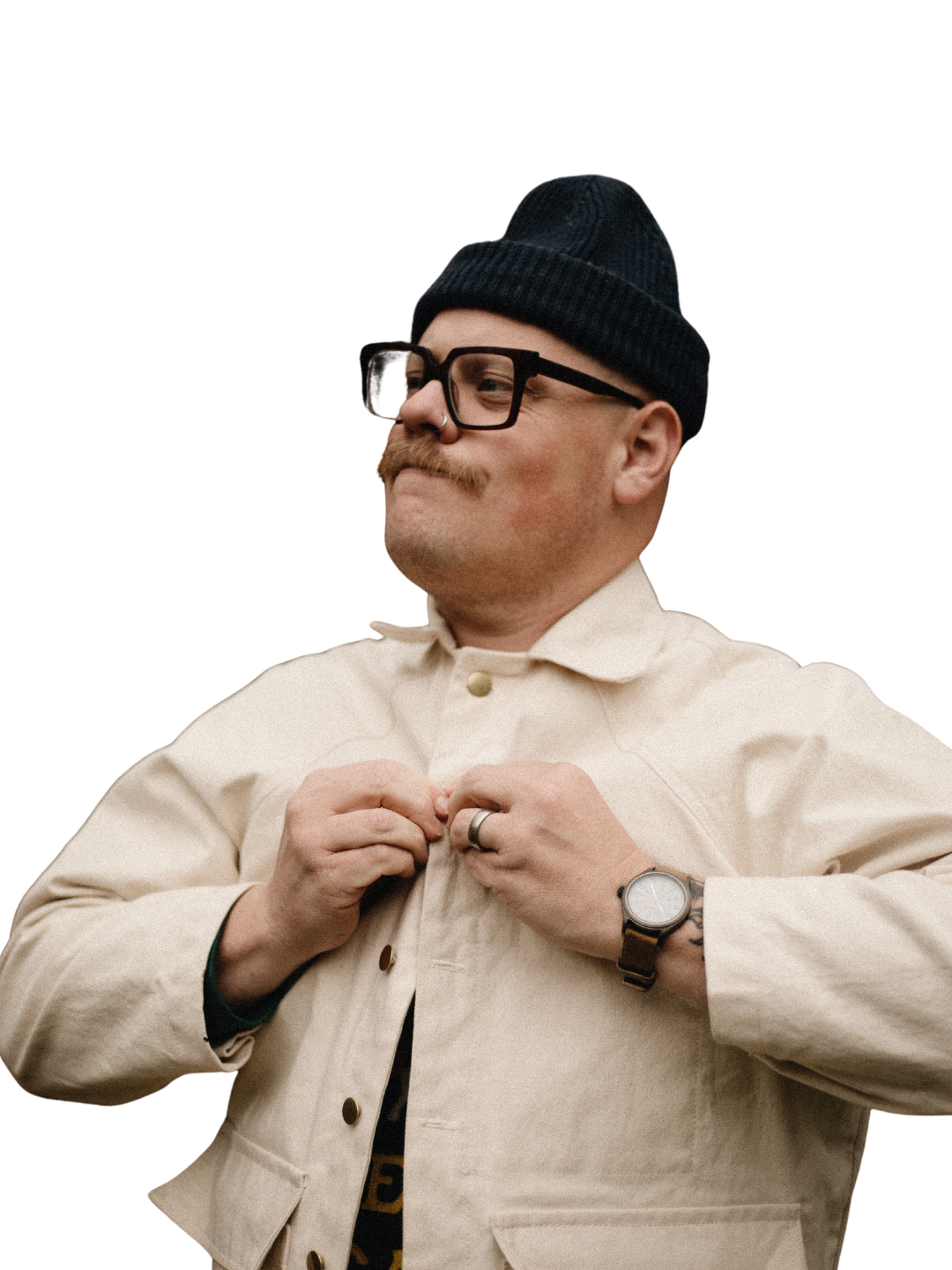 Man in black beanie and glasses with a handlebar mustache buttons a cream work jacket while wearing a wristwatch against a white background.