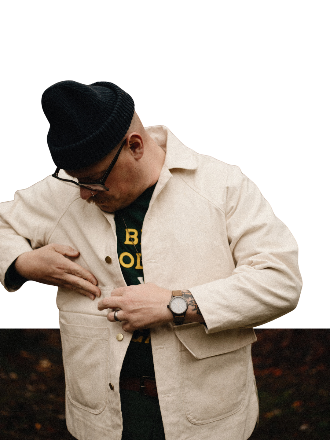 Man in a black knit beanie, glasses and cream chore jacket over a graphic tee adjusts the buttons while wearing a wristwatch outdoors.