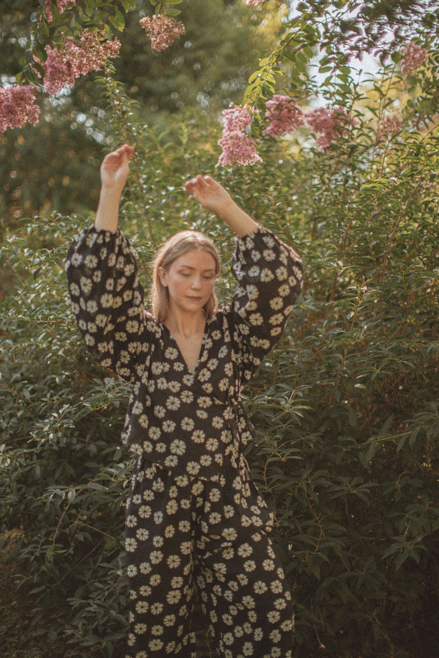 Woman in floral black dress with arms raised among blooming pink flowers and lush green bushes outdoors.