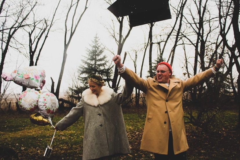 Couple celebrating outdoors in autumn park, woman holding balloons, man with arms raised, wearing coats.