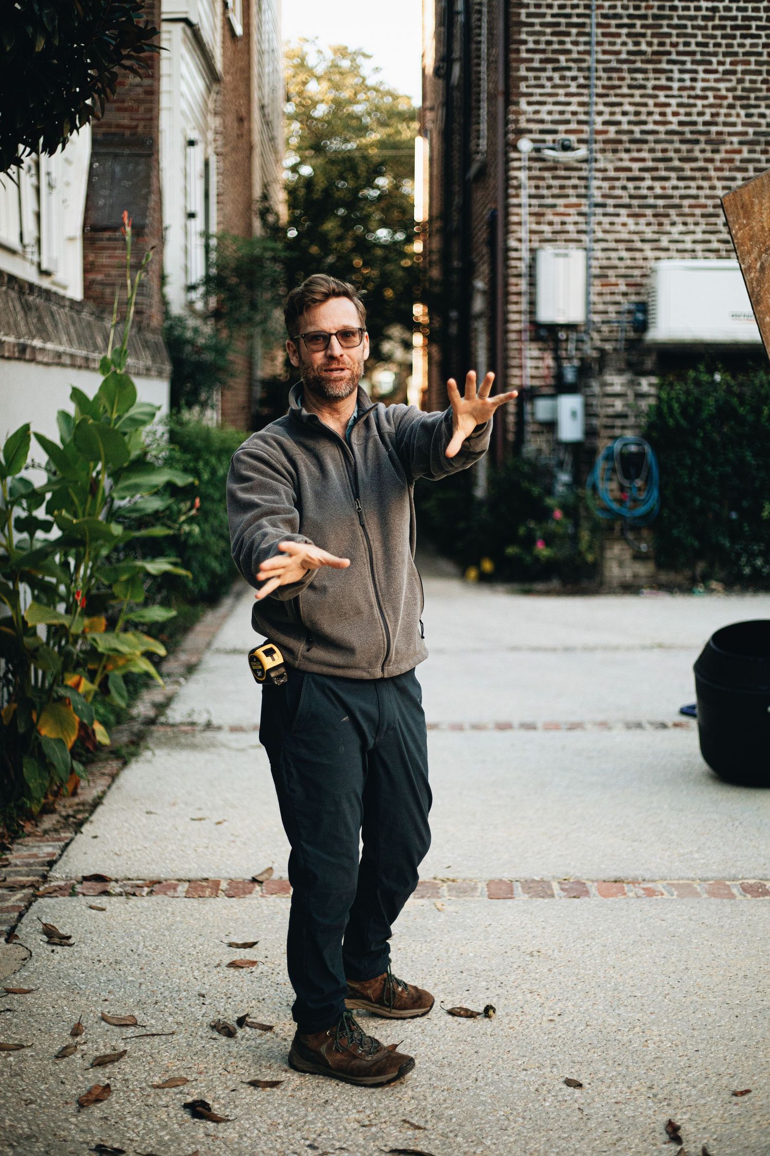 Man in glasses and a gray fleece extends both arms outward while standing on a brick-lined alley path beside green plants.