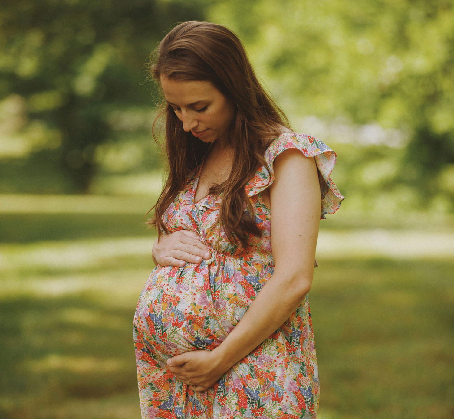 Pregnant woman in floral dress cradling baby bump outdoors in lush green park during maternity photo session.