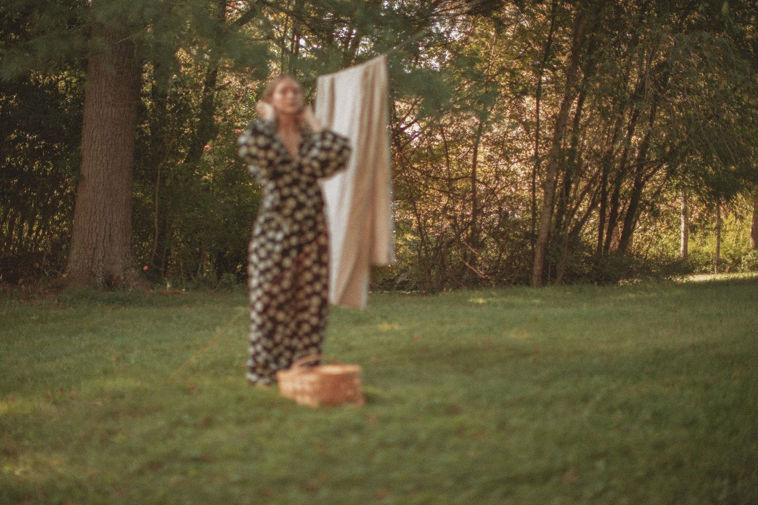 Woman in floral dress hanging laundry on clothesline in lush green backyard during golden hour.