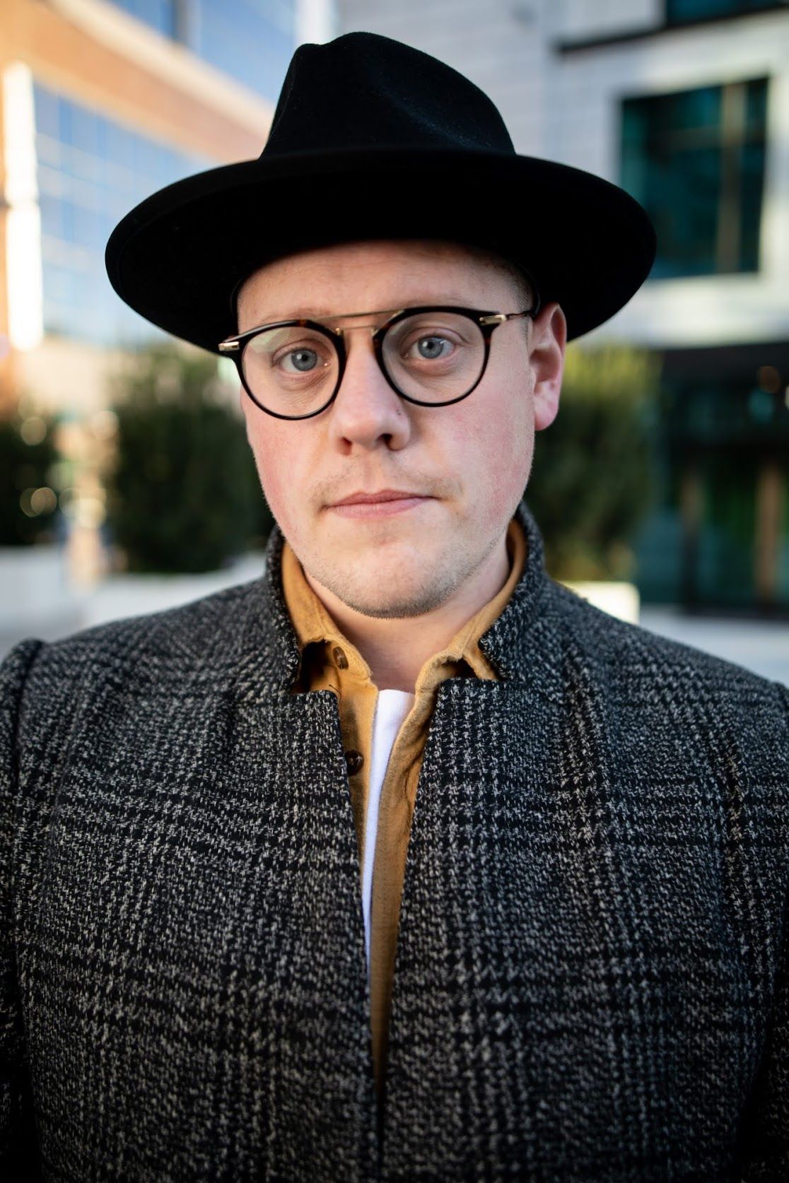 Man in black wide-brim hat and round glasses wearing plaid coat outdoors in urban setting.