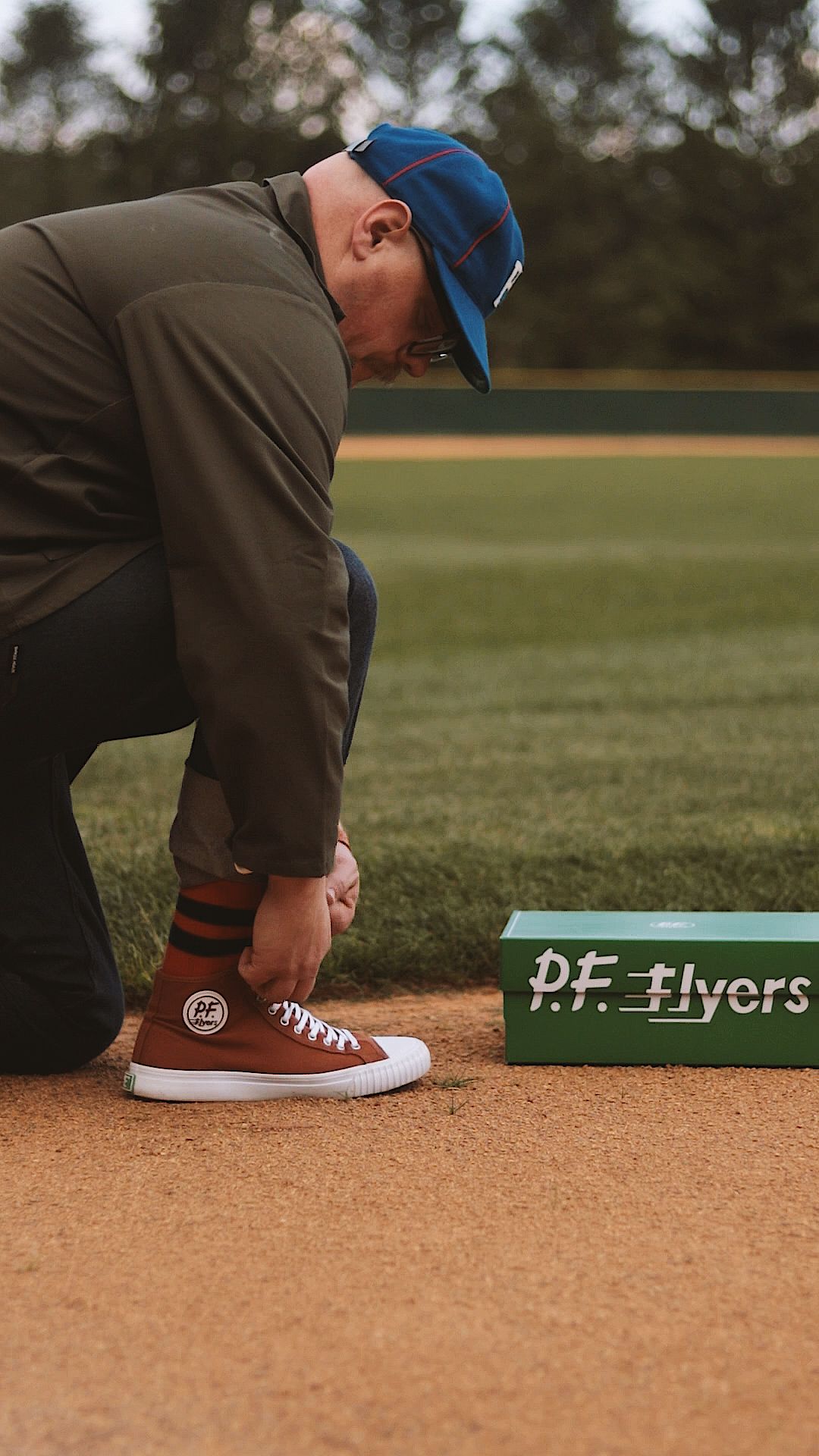Person tying P.F. Flyers sneakers on a baseball field dirt infield, wearing a blue cap and olive jacket.