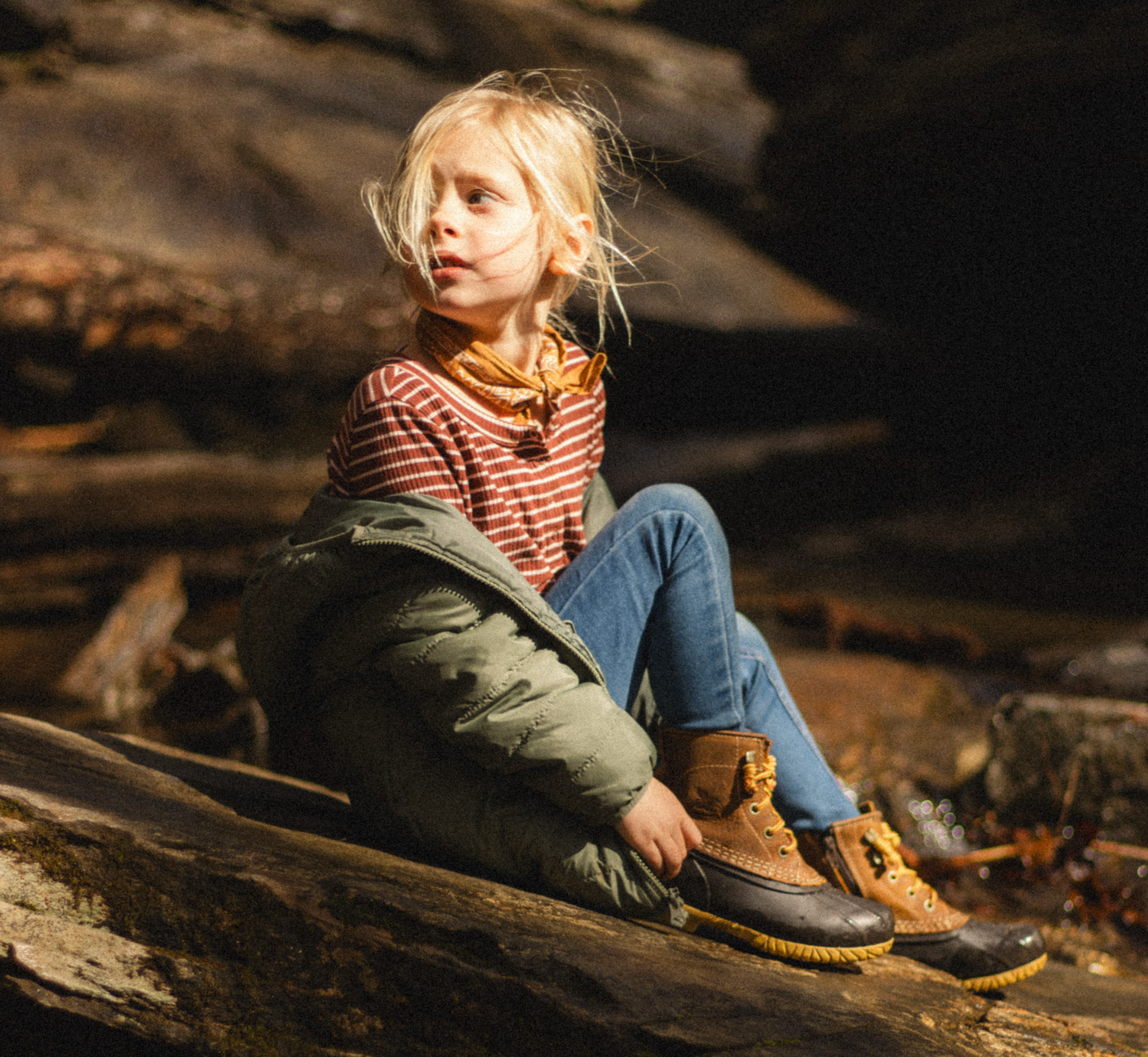 A young child with blonde hair sits on rocks outdoors, wearing striped shirt, jeans, and brown duck boots.