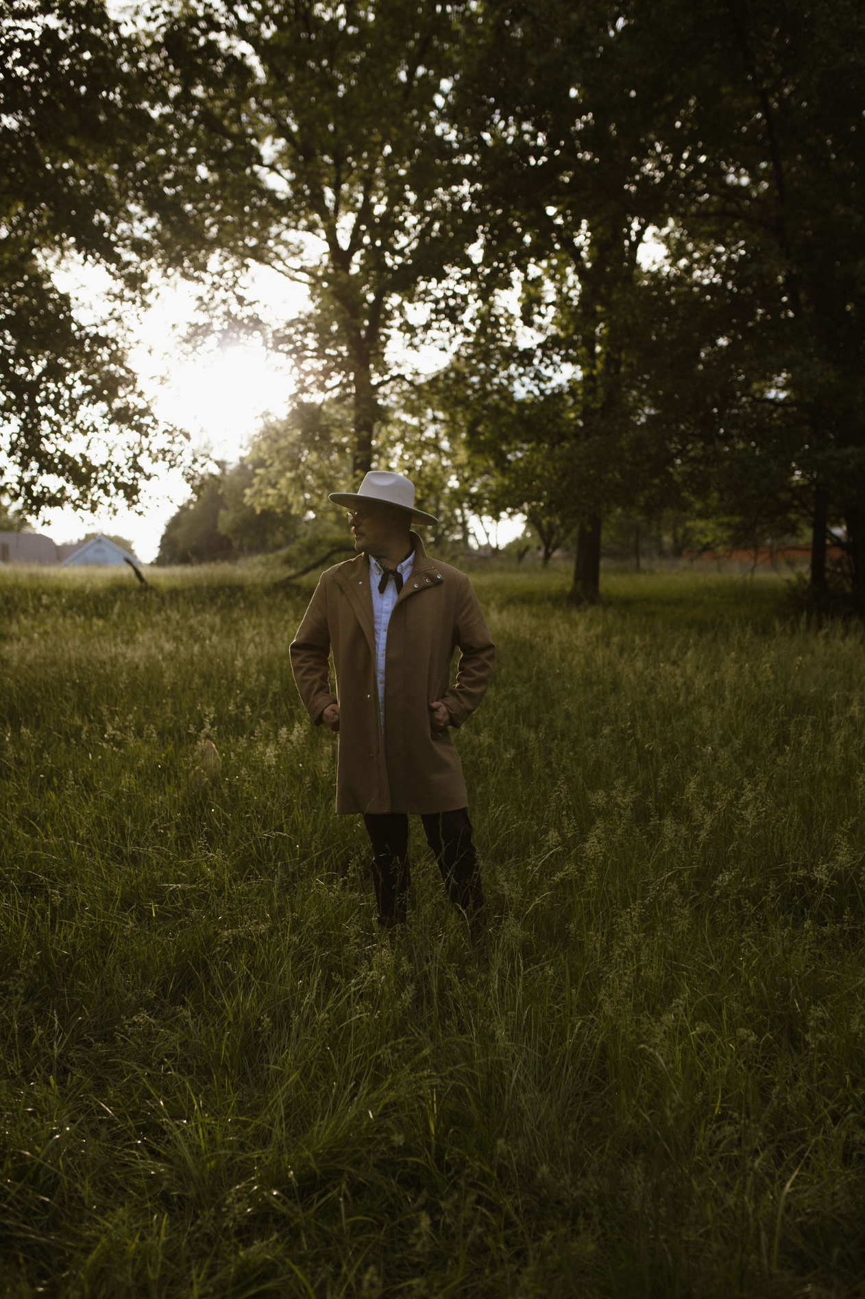 Man in brown coat and cowboy hat standing in lush green field with sunlit trees in background.