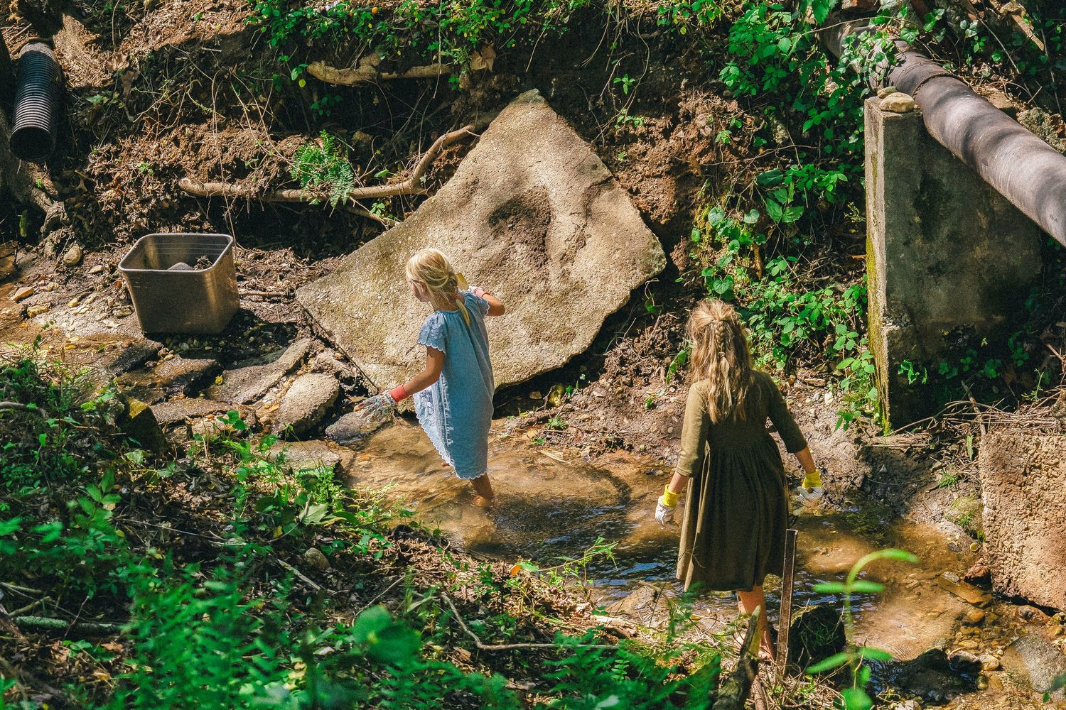 Two young girls exploring a rocky stream in a lush green forest setting on a sunny day.