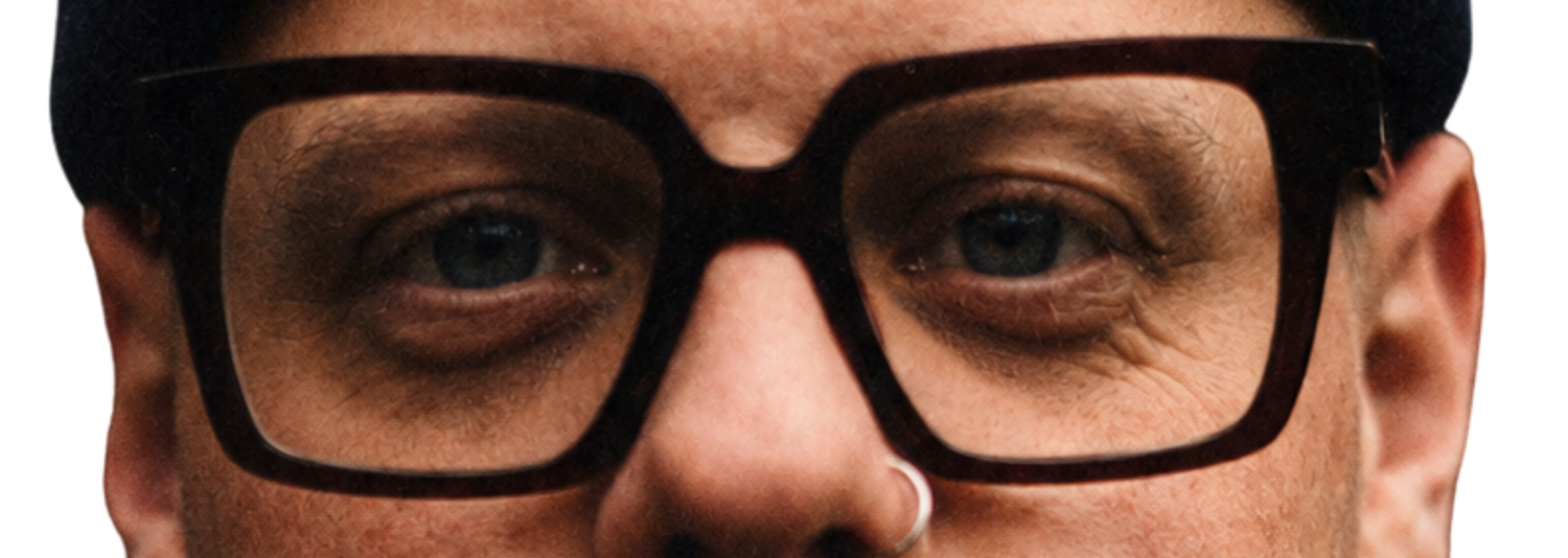 Close-up of a person's eyes behind thick black rectangular glasses, with a small silver nose stud visible.