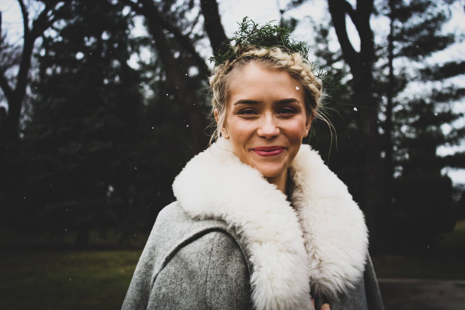 A smiling young woman wearing a floral crown and gray coat with white fur collar stands outdoors among dark trees.