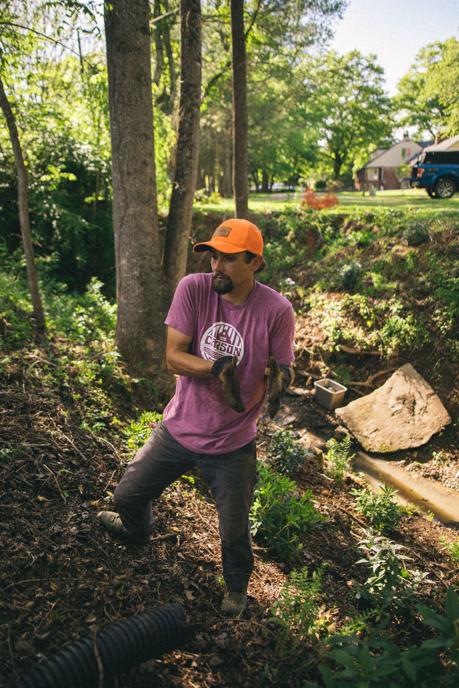 Man in orange cap and pink shirt doing outdoor landscaping work in a wooded backyard with drainage pipes.