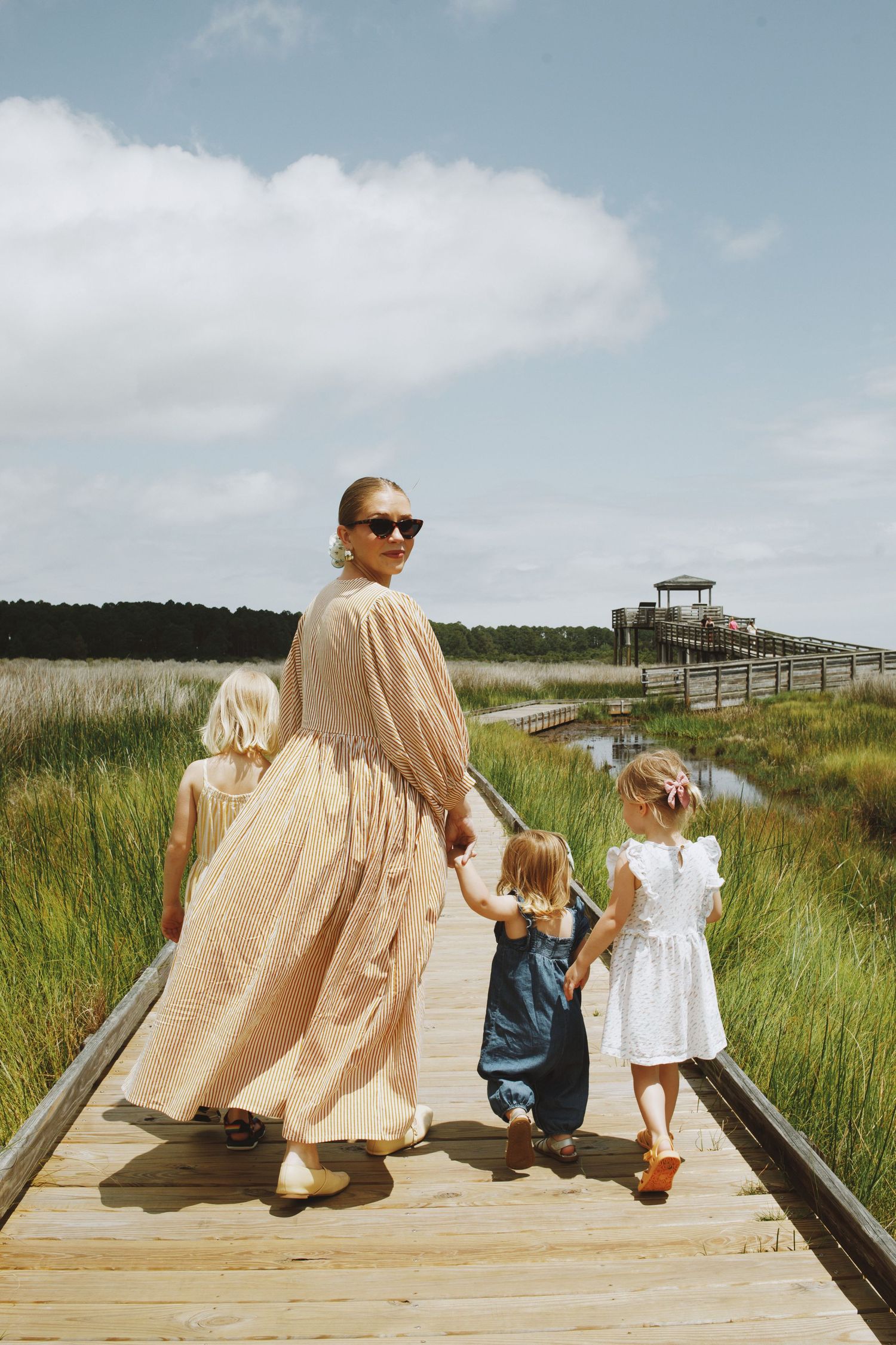 A woman in a beige dress walks with three young girls on a wooden boardwalk through a coastal marsh.