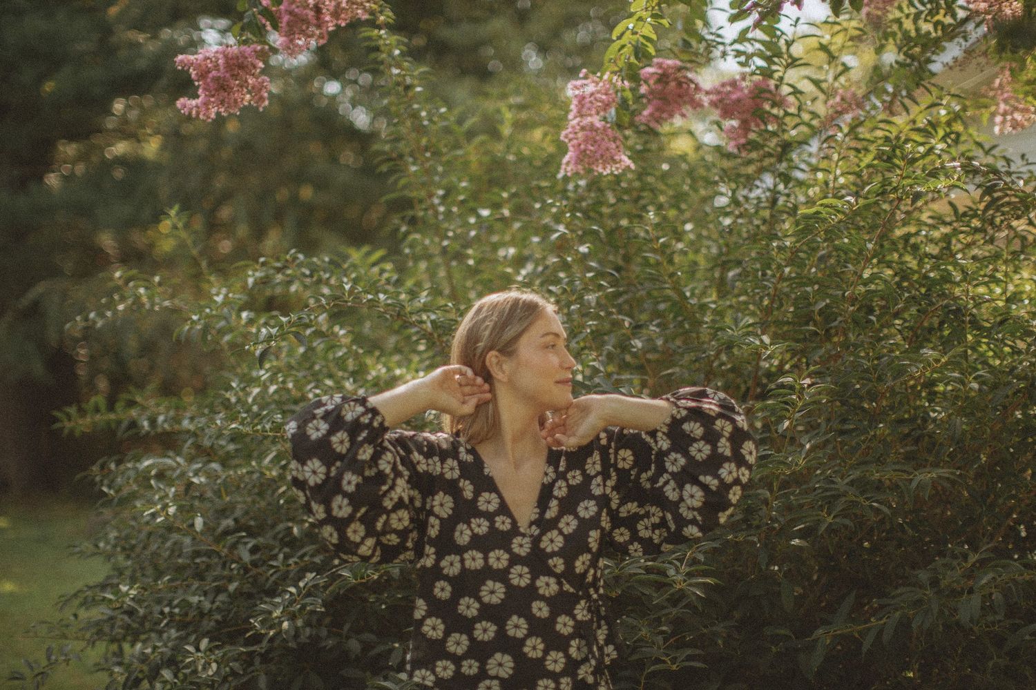 Woman in floral dress posing gracefully among lush green bushes and pink blooming flowers in a sunlit garden.