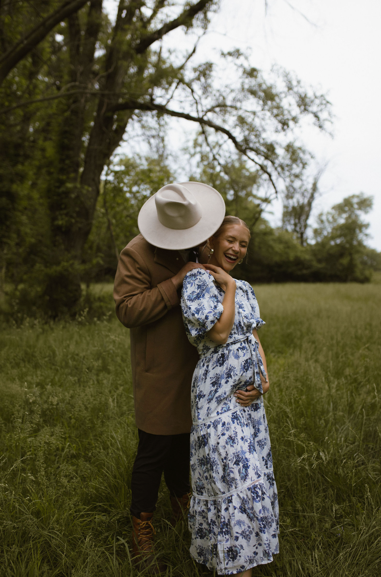 A couple embraces in a green meadow, the woman in a floral dress holding her baby bump, laughing joyfully.