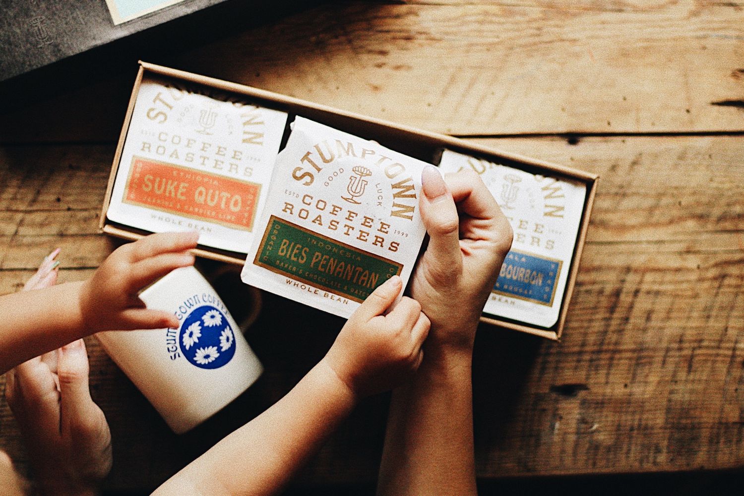 Hands holding coffee sample boxes and cupping cards on a wooden surface next to a coffee tasting spoon.