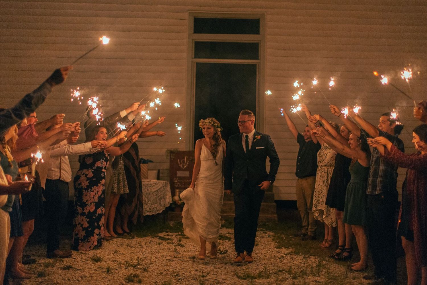 Bride and groom walk through sparkler tunnel with wedding guests at night outside white building.
