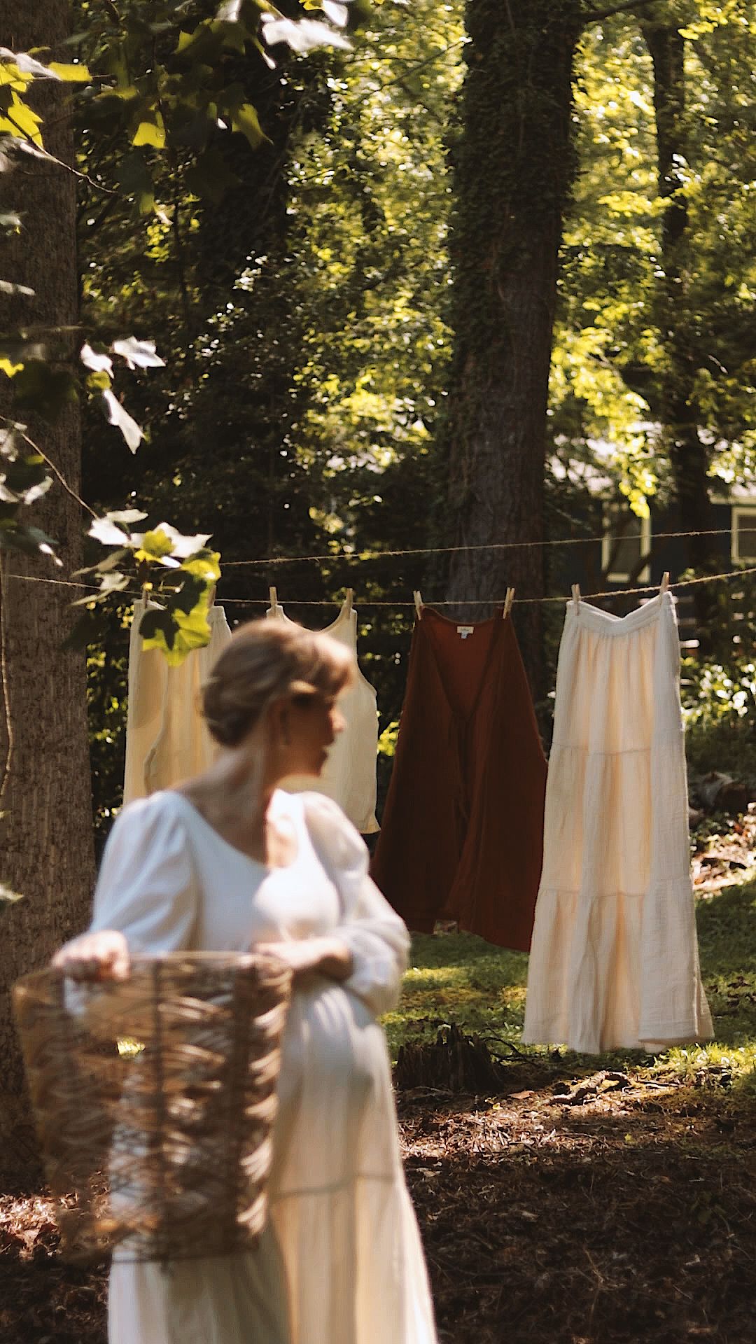 Woman in white dress carrying wicker basket near clothesline with hanging garments in sunlit garden.