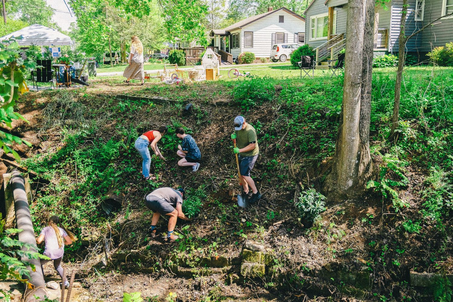 Group of volunteers digging and landscaping a backyard garden on a sunny day surrounded by trees.