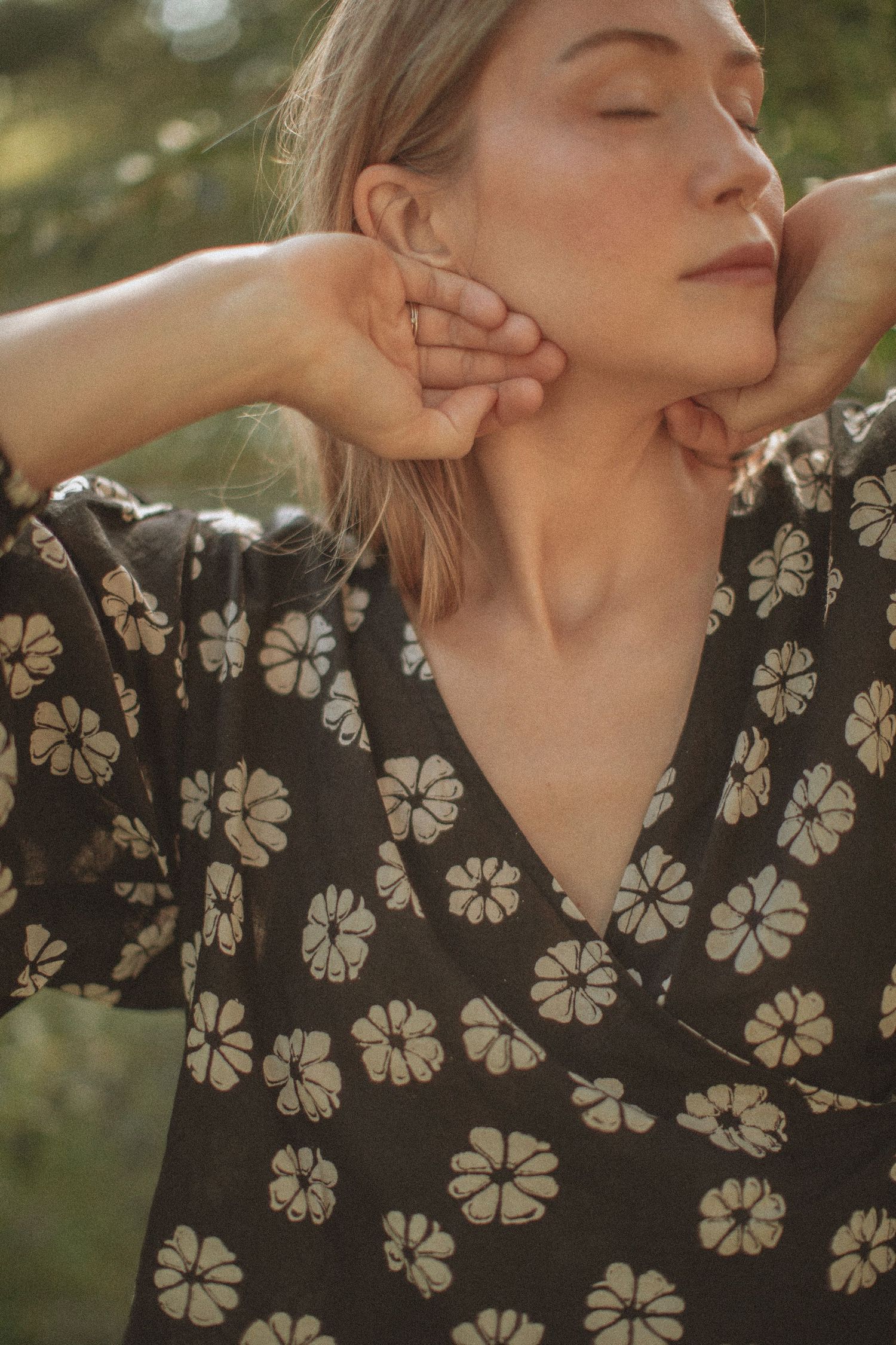 Woman in black floral wrap dress poses outdoors with hands raised to neck, eyes closed in warm golden light.