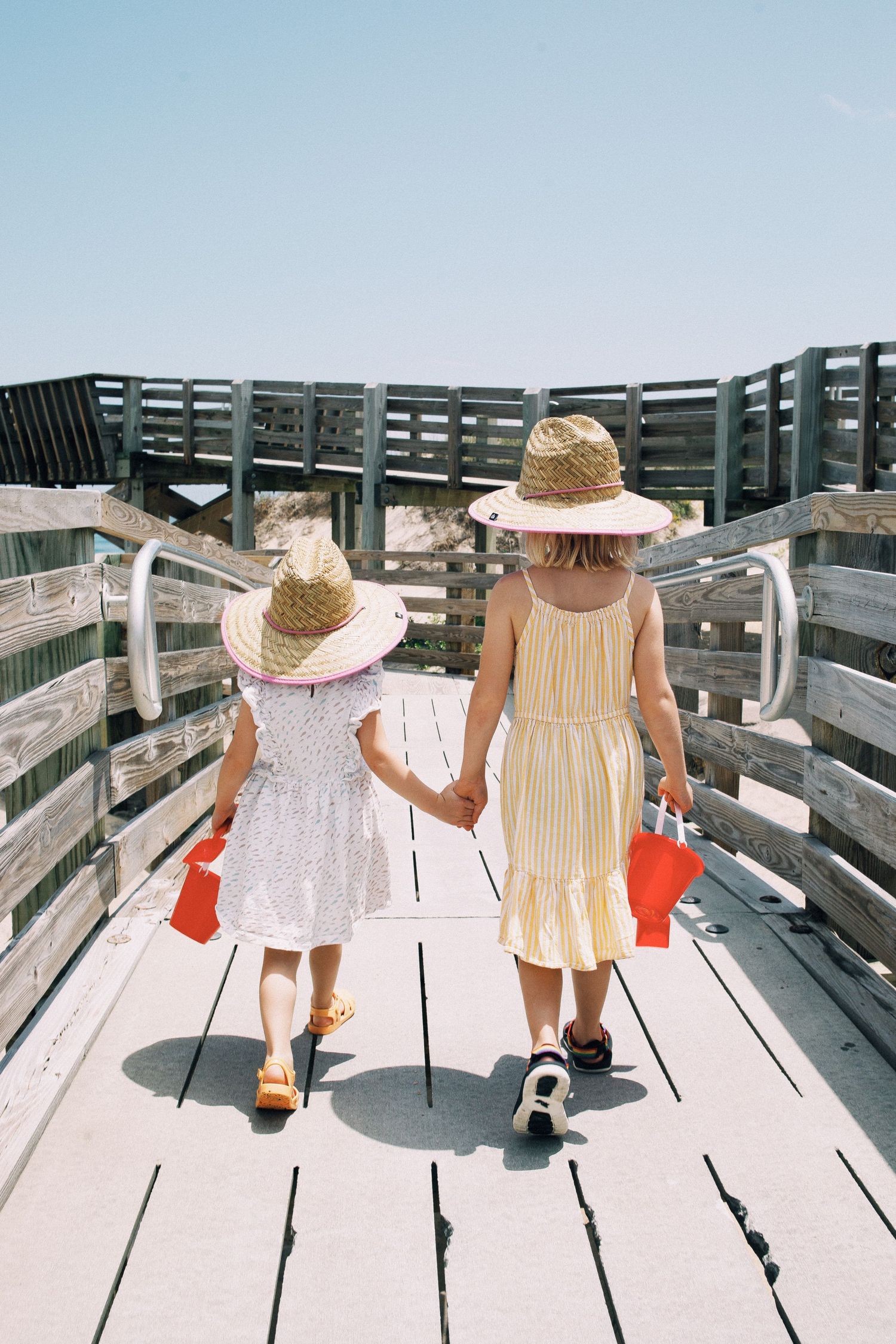 Two young girls in straw hats and summer dresses hold hands walking on a wooden boardwalk to the beach.