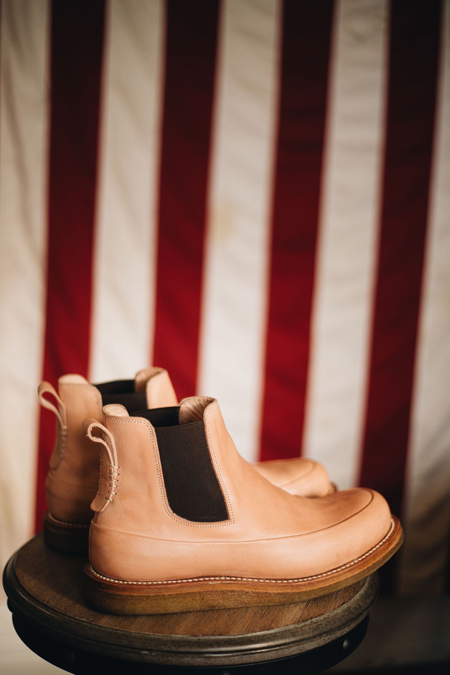 Tan leather Chelsea boots displayed on a wooden stand against a red and white striped American flag background.