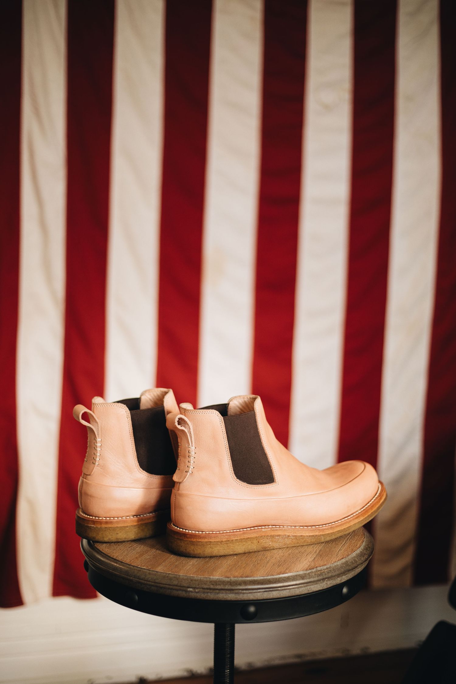 Tan leather Chelsea boots displayed on a wooden stand against a red and white striped American flag backdrop.